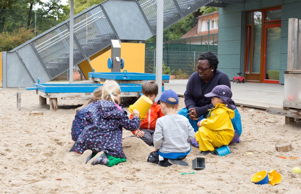 Kinder spielen im Sandkasten. Eine Erzieherin sitzt neben den Kindern.